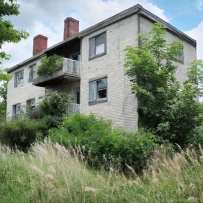 AI generated image of a small apartment building with overgrown bushes and long grass.